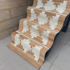 May include: A woven jute stair runner featuring a repeating geometric pattern in natural tan and off-white. The runner is draped over a set of stairs, showcasing a textured, fibrous material with a consistent weave and a clean, finished edge.