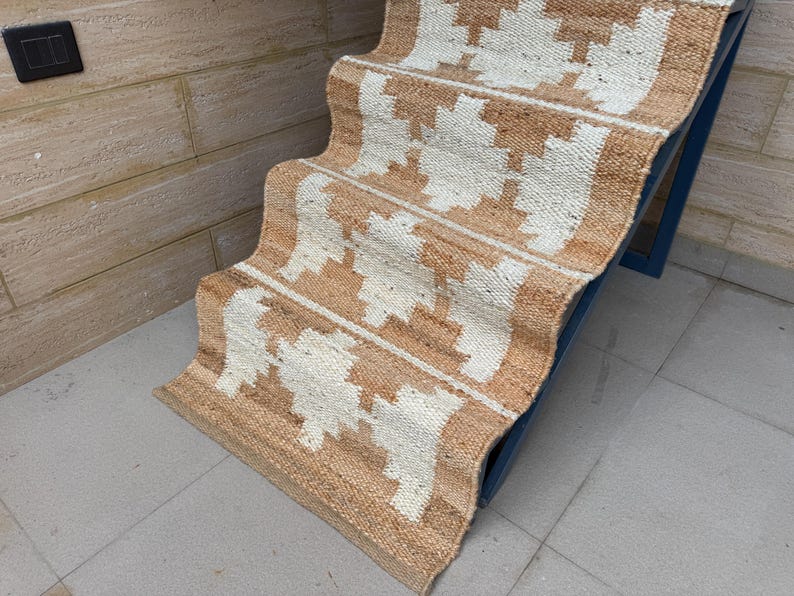 May include: A woven jute stair runner featuring a repeating geometric pattern in natural tan and off-white. The runner is draped over a set of stairs, showcasing a textured, fibrous material with a consistent weave and a clean, finished edge.