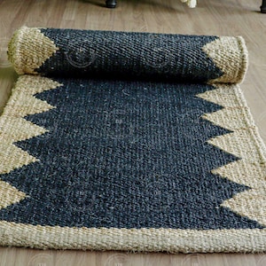 Op de afbeelding: Een opgerolde, rechthoekige vloerkleed met een donkerblauwe kern en een natuurlijke beige rand. De rand heeft een zigzagpatroon. Het vloerkleed is gemaakt van geweven materiaal en ligt op een houten vloer.