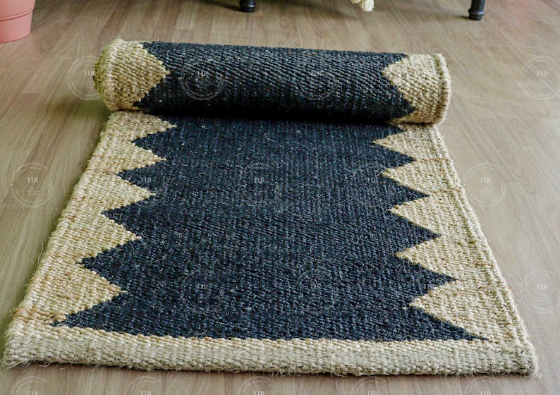Op de afbeelding: Een opgerolde, rechthoekige vloerkleed met een donkerblauwe kern en een natuurlijke beige rand. De rand heeft een zigzagpatroon. Het vloerkleed is gemaakt van geweven materiaal en ligt op een houten vloer.