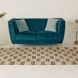 May include: A teal velvet sofa with gold legs and two light blue pillows. A beige rug with a black geometric pattern is in front of the sofa. A small side table and a white cabinet are on the right, and a potted plant is on the left.