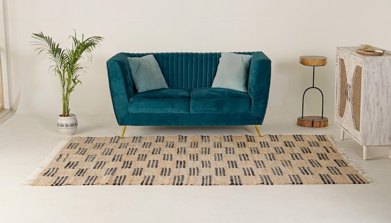 May include: A teal velvet sofa with gold legs and two light blue pillows. A beige rug with a black geometric pattern is in front of the sofa. A small side table and a white cabinet are on the right, and a potted plant is on the left.