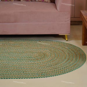 Puede incluir: Alfombra ovalada de yute con un dise&ntilde;o trenzado. La alfombra presenta un color marr&oacute;n natural con un borde verde. La alfombra est&aacute; colocada sobre un suelo de color claro, con un sof&aacute; rosa y una mesa de madera en el fondo.