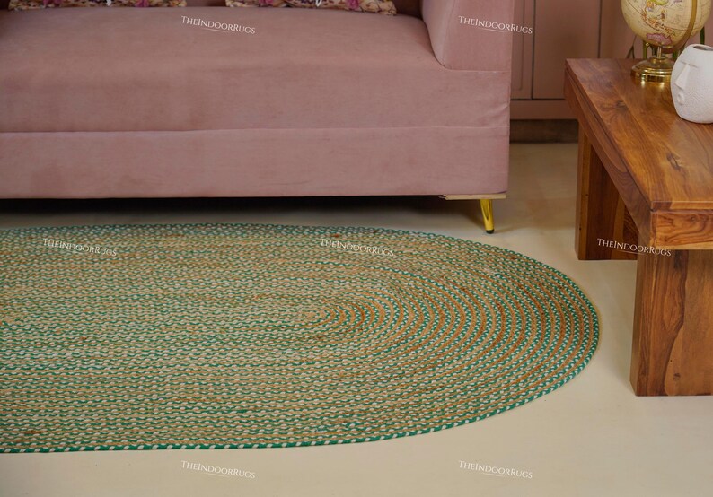 Puede incluir: Alfombra ovalada de yute con un dise&ntilde;o trenzado. La alfombra presenta un color marr&oacute;n natural con un borde verde. La alfombra est&aacute; colocada sobre un suelo de color claro, con un sof&aacute; rosa y una mesa de madera en el fondo.