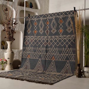 Può includere: Un tappeto grigio scuro con motivi geometrici bianchi e arancioni. Il tappeto è appeso a un'asta di metallo. Il design include motivi a diamante, triangolo e linea. Il tappeto ha un bordo sfrangiato ed è esposto in una stanza con vasi e piante decorative.