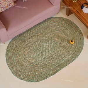 Puede incluir: Una alfombra ovalada con textura tejida en tonos verdes y beige. La alfombra est&aacute; sobre un suelo claro, con un sof&aacute; rosa y una mesa de madera al fondo. Un peque&ntilde;o jarr&oacute;n amarillo con vegetaci&oacute;n est&aacute; sobre la alfombra. La alfombra mide unos 200 cm de largo.