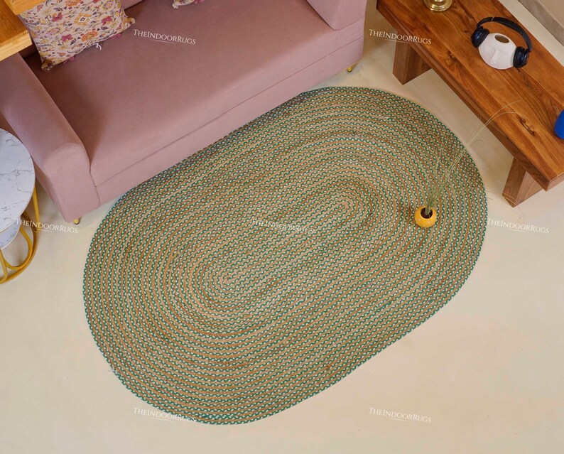 Puede incluir: Una alfombra ovalada con textura tejida en tonos verdes y beige. La alfombra est&aacute; sobre un suelo claro, con un sof&aacute; rosa y una mesa de madera al fondo. Un peque&ntilde;o jarr&oacute;n amarillo con vegetaci&oacute;n est&aacute; sobre la alfombra. La alfombra mide unos 200 cm de largo.