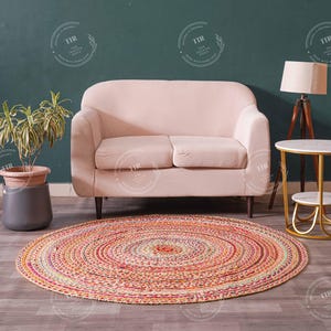 K&ouml;nnte beinhalten: Ein runder, mehrfarbiger geflochtener Teppich mit konzentrischen Kreisen liegt auf einem Holzboden. Im Hintergrund befinden sich ein hellrosa Sofa, ein Beistelltisch mit Marmorplatte und eine Stehlampe. Links ist eine Topfpflanze.