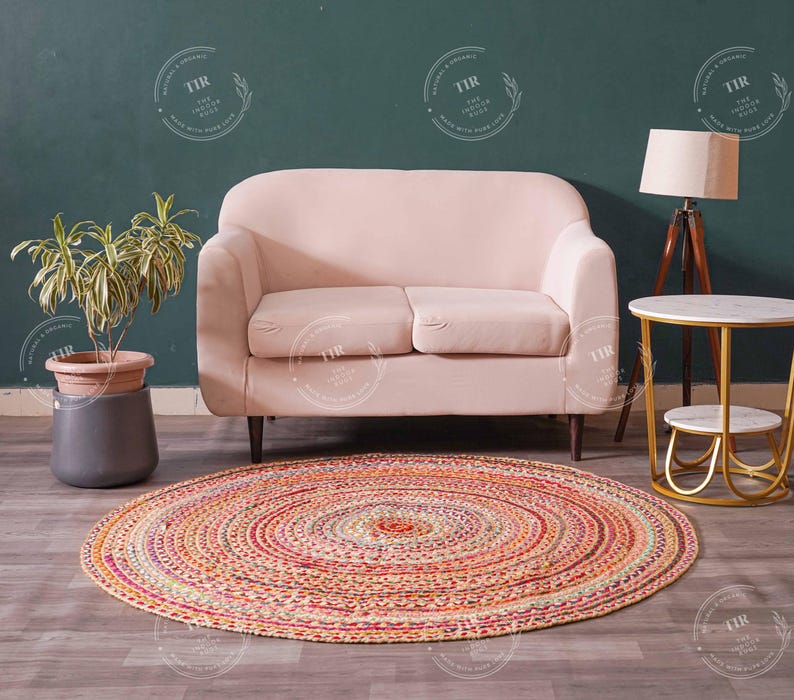 K&ouml;nnte beinhalten: Ein runder, mehrfarbiger geflochtener Teppich mit konzentrischen Kreisen liegt auf einem Holzboden. Im Hintergrund befinden sich ein hellrosa Sofa, ein Beistelltisch mit Marmorplatte und eine Stehlampe. Links ist eine Topfpflanze.