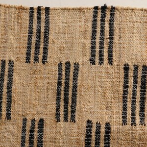 May include: Close-up of a woven rug with a natural beige background and vertical black stripes. The rug has a textured surface and a fringe detail on the right side. The design is simple and geometric.
