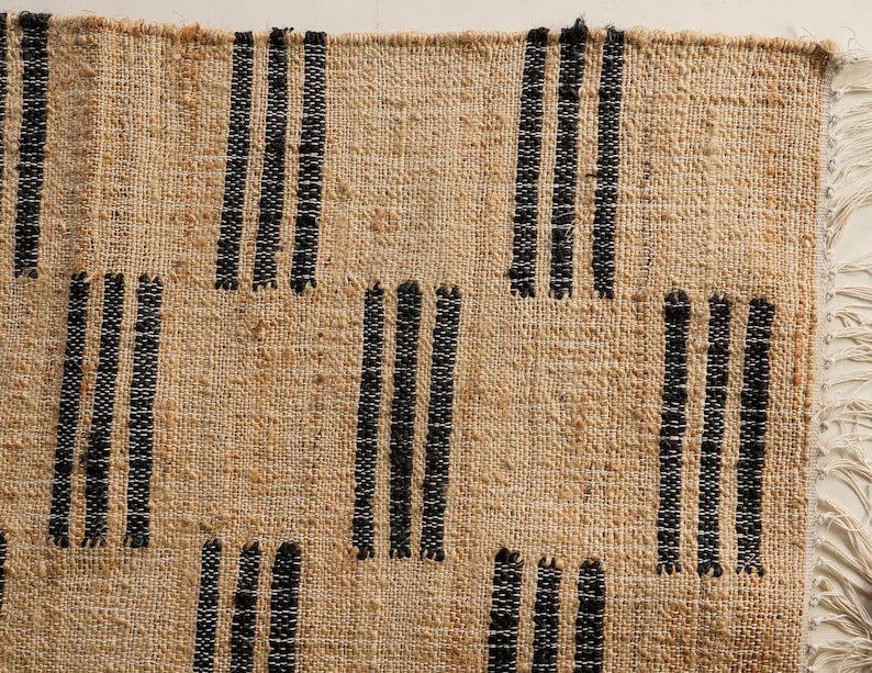 May include: Close-up of a woven rug with a natural beige background and vertical black stripes. The rug has a textured surface and a fringe detail on the right side. The design is simple and geometric.