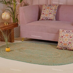 Puede incluir: Una alfombra ovalada de yute trenzado con borde verde sobre un suelo claro. Detr&aacute;s de la alfombra hay un sof&aacute; rosa con patas doradas y cojines estampados. A la izquierda, una mesa de madera con un globo terr&aacute;queo y una planta.