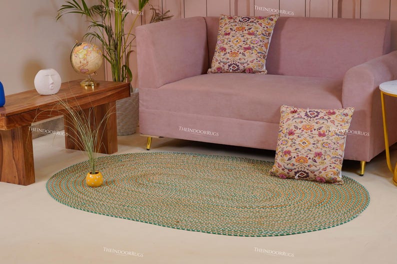 Puede incluir: Una alfombra ovalada de yute trenzado con borde verde sobre un suelo claro. Detr&aacute;s de la alfombra hay un sof&aacute; rosa con patas doradas y cojines estampados. A la izquierda, una mesa de madera con un globo terr&aacute;queo y una planta.