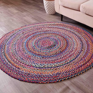 K&ouml;nnte beinhalten: Ein runder geflochtener Teppich mit einem lebendigen, mehrfarbigen Design. Der Teppich weist konzentrische Kreise aus verflochtenen F&auml;den in Rot-, Blau-, Orange- und Gr&uuml;nt&ouml;nen auf. Der Teppich liegt auf einem Holzboden.