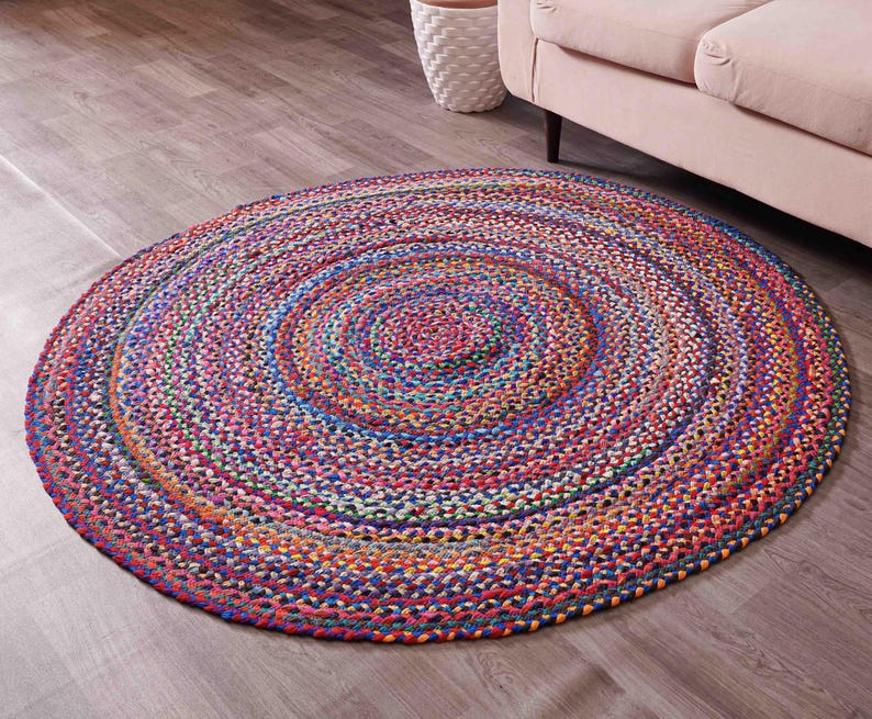 K&ouml;nnte beinhalten: Ein runder geflochtener Teppich mit einem lebendigen, mehrfarbigen Design. Der Teppich weist konzentrische Kreise aus verflochtenen F&auml;den in Rot-, Blau-, Orange- und Gr&uuml;nt&ouml;nen auf. Der Teppich liegt auf einem Holzboden.