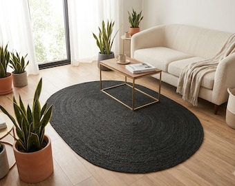 May include: An oval-shaped area rug with a dark charcoal black braided texture. The rug is positioned on a light-colored wooden floor in a room featuring a cream-colored sofa, a wooden coffee table with a gold frame, and several potted snake plants.