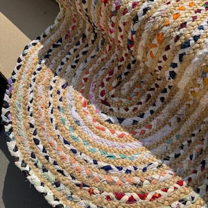 Könnte beinhalten: Runder geflochtener Teppich mit Spiralmuster. Der Teppich zeigt eine Mischung aus naturfarbenem Beige, Weiß und farbigen Stoffstreifen in einem sich wiederholenden Muster. Der Teppich besteht aus gewebten Materialien.