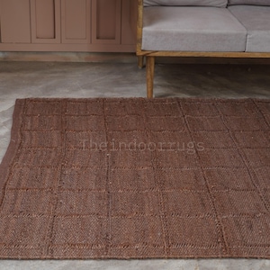 May include: A rectangular brown area rug featuring a grid pattern of woven squares. The rug has a textured surface with visible fiber construction and a finished edge border. The text Theindoorrugs is printed in the center of the rug.