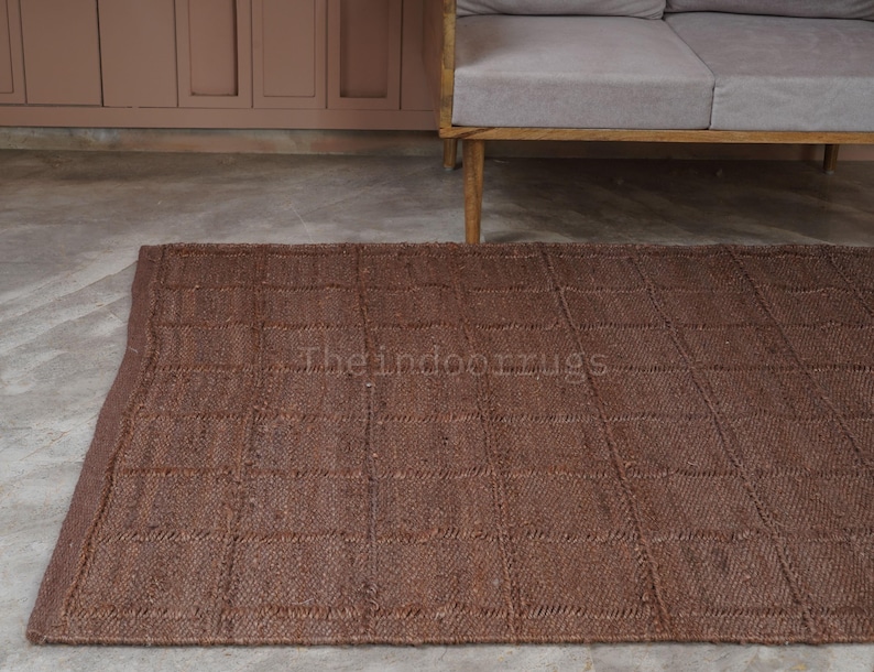 May include: A rectangular brown area rug featuring a grid pattern of woven squares. The rug has a textured surface with visible fiber construction and a finished edge border. The text Theindoorrugs is printed in the center of the rug.