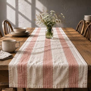 Pode incluir: Um caminho de mesa retangular com uma base de cor creme e riscas verticais de cor terracota. O tecido tem uma textura tecida visível e está colocado na horizontal sobre o centro de uma mesa de jantar de madeira, de uma ponta à outra.