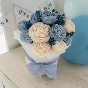 Könnte beinhalten: Ein Blumenstrauß aus geformten cremefarbenen und staubblauen blütenförmigen Seifen in einem runden weißen Behälter. Das Arrangement ist in weißes Seidenpapier eingewickelt und mit einem staubblauen Band gebunden. Kleine dunkelblaue Trockenblumen.