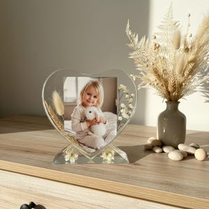 Può includere: Una cornice portafoto in resina trasparente a forma di cuore contenente la fotografia di un bambino che tiene in mano un coniglietto di peluche bianco. La cornice è decorata con erbe beige essiccate e piccoli fiori bianchi su base triangolare.