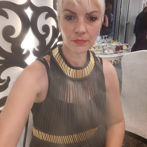 May include: A woman wearing a black mesh dress with a gold chain detail around the neck and waist. The dress has a halter neckline and is sleeveless.