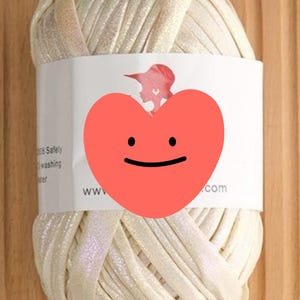 May include: A ball of cream-colored yarn with an iridescent glitter finish. The yarn is wrapped in a white paper label featuring a red heart graphic, a silhouette of a person, and text that reads Cream with iridescent glitter.