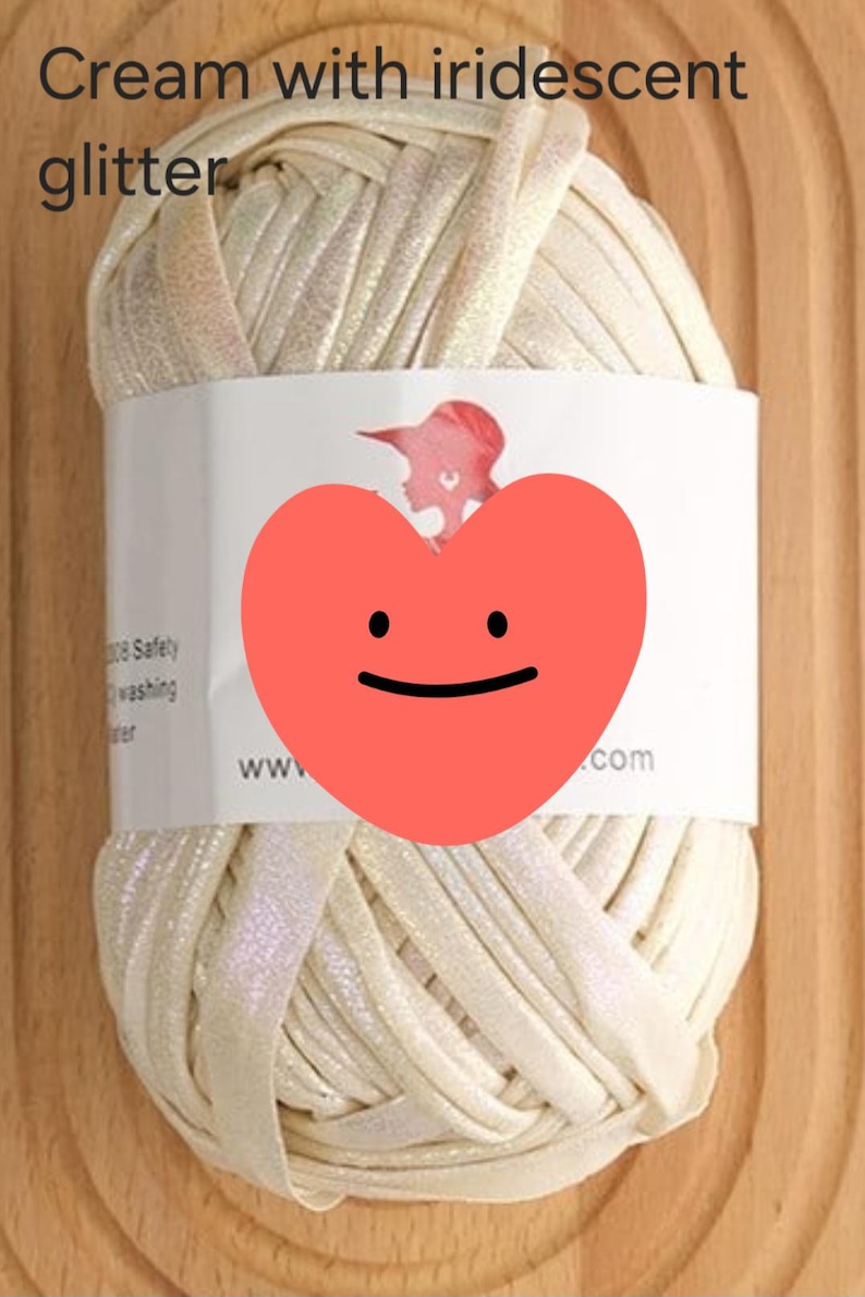 May include: A ball of cream-colored yarn with an iridescent glitter finish. The yarn is wrapped in a white paper label featuring a red heart graphic, a silhouette of a person, and text that reads Cream with iridescent glitter.