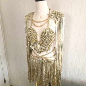May include: A metallic gold two-piece outfit featuring a halter-style crop top and a matching skirt, both adorned with long, shimmering fringe. The set includes decorative gold chain accents at the neckline and waist, with integrated shoulder capes.