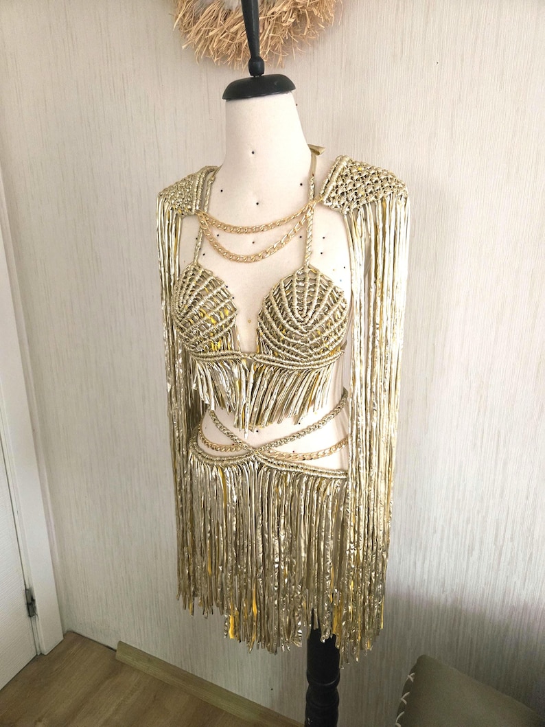 May include: A metallic gold two-piece outfit featuring a halter-style crop top and a matching skirt, both adorned with long, shimmering fringe. The set includes decorative gold chain accents at the neckline and waist, with integrated shoulder capes.