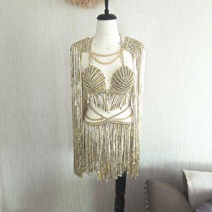 May include: A metallic gold macrame two-piece set featuring a shell-shaped bralette top and a matching fringe skirt. The set includes long fringe shoulder drapes and thin gold chain accents connecting the top and skirt, displayed on a mannequin.