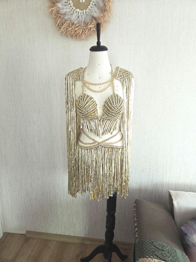 May include: A metallic gold macrame two-piece set featuring a shell-shaped bralette top and a matching fringe skirt. The set includes long fringe shoulder drapes and thin gold chain accents connecting the top and skirt, displayed on a mannequin.