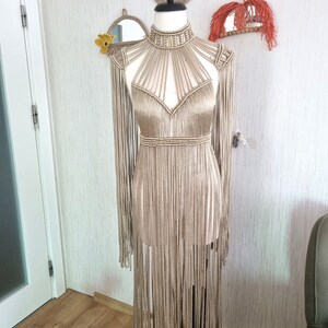 Puede incluir: Un vestido largo de crochet beige con escote halter y detalles de flecos. El vestido est&aacute; en un maniqu&iacute; y tiene un escote alto.