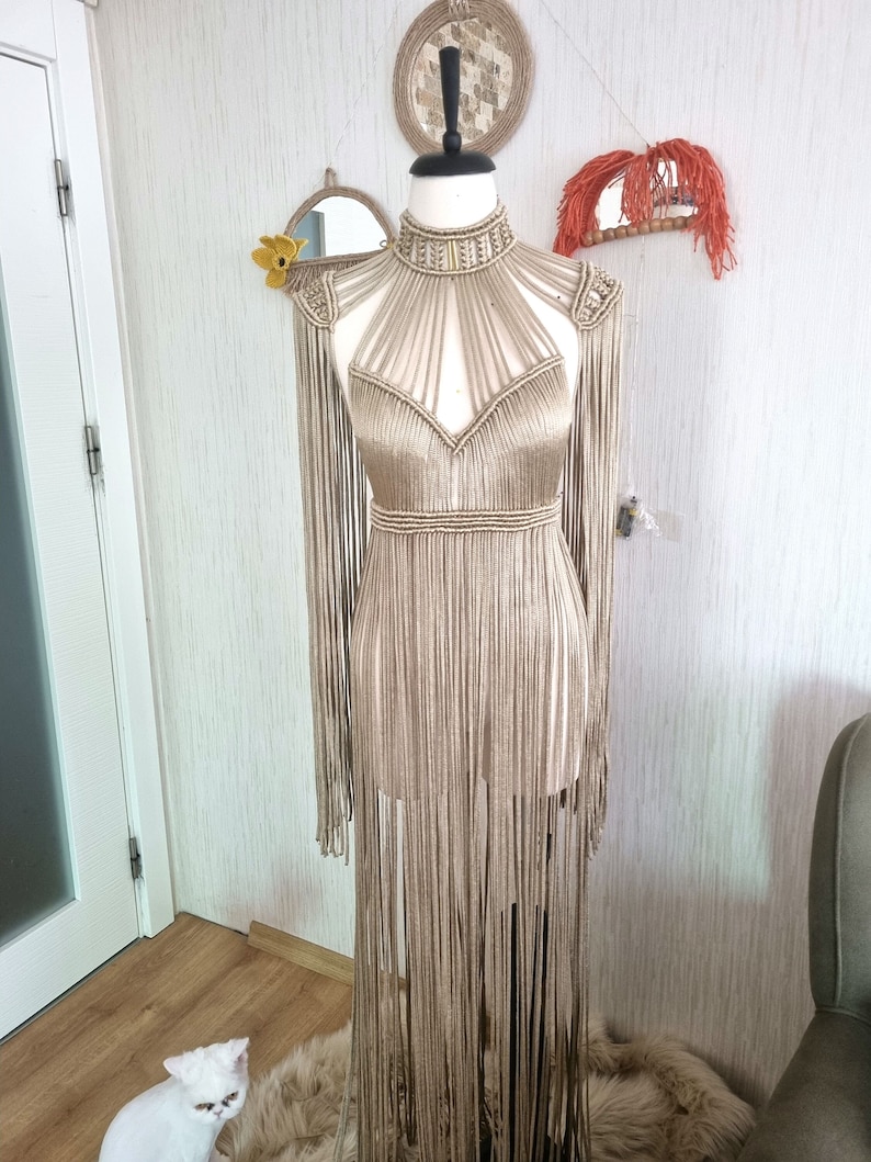 Puede incluir: Un vestido largo de crochet beige con escote halter y detalles de flecos. El vestido est&aacute; en un maniqu&iacute; y tiene un escote alto.