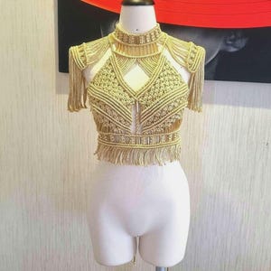 May include: A gold macrame top with fringe detailing. The top is on a white dress form.