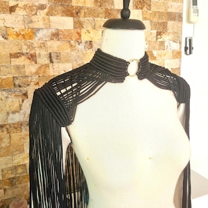 May include: Black macrame shoulder piece with fringe and a gold ring detail. The piece is worn on a white mannequin.