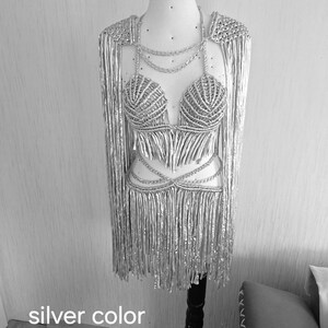 May include: A metallic silver two-piece set featuring a macrame-style bralette with a V-neckline and a matching skirt, both adorned with long, flowing fringe. The set includes integrated shoulder capes and chain-like straps in silver color.