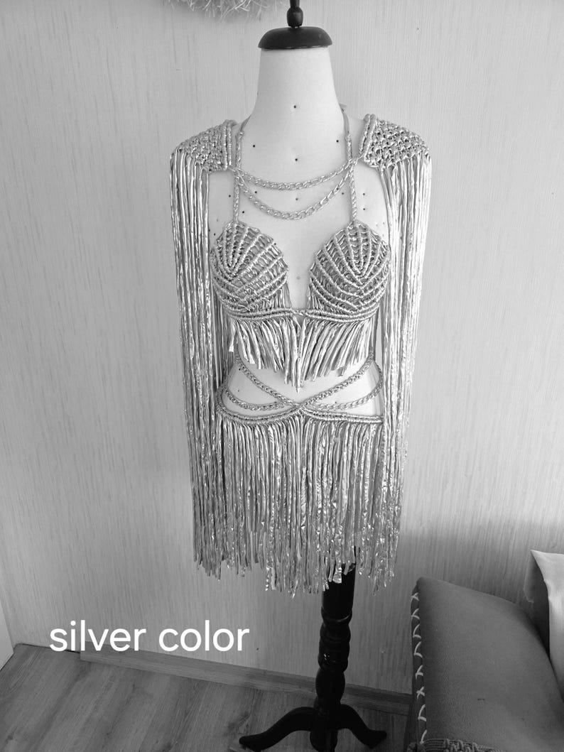 May include: A metallic silver two-piece set featuring a macrame-style bralette with a V-neckline and a matching skirt, both adorned with long, flowing fringe. The set includes integrated shoulder capes and chain-like straps in silver color.