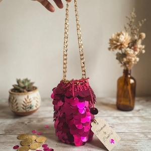 May include: A cylindrical pouch covered in vibrant fuchsia sequins with a gold-tone metal chain strap. The top features a drawstring closure made of matching fuchsia yarn. A beige paper tag attached to the side reads Handmade by Creators.