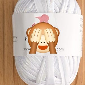 May include: A ball of white yarn with a subtle glitter finish, wrapped in a white paper band. The band features partial black text and a graphic of a monkey covering its eyes. The yarn is composed of thick, flat strands of white material.