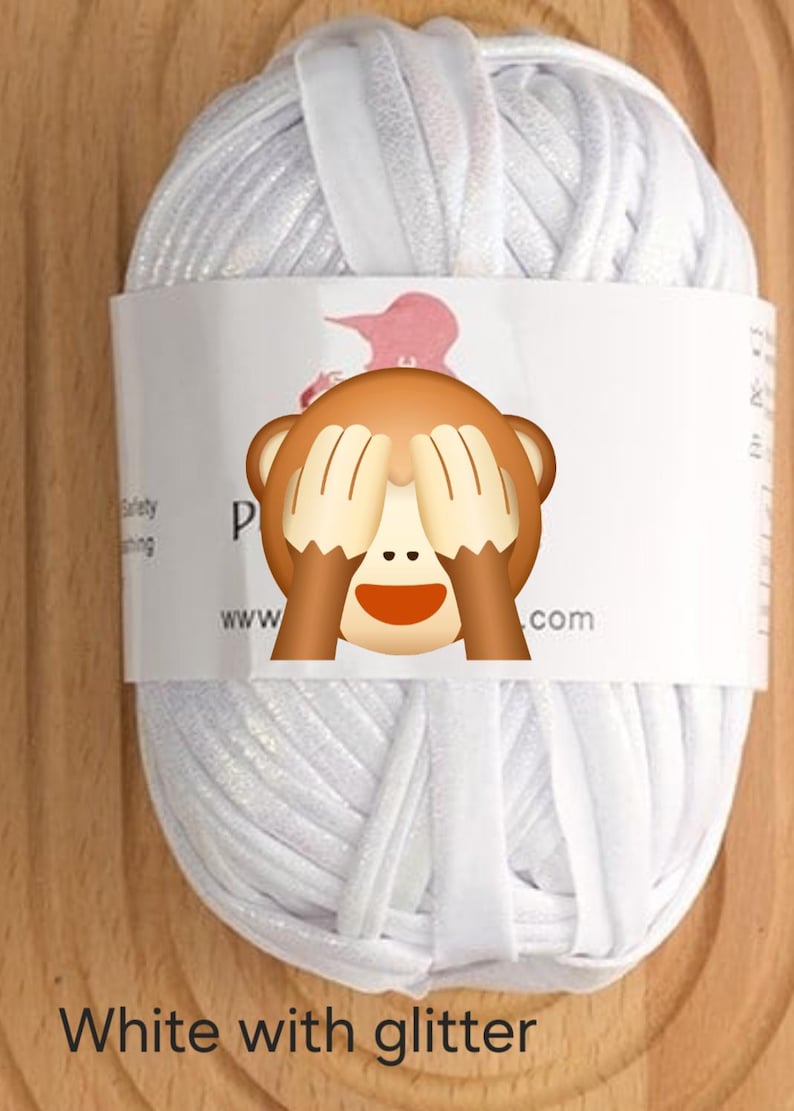 May include: A ball of white yarn with a subtle glitter finish, wrapped in a white paper band. The band features partial black text and a graphic of a monkey covering its eyes. The yarn is composed of thick, flat strands of white material.
