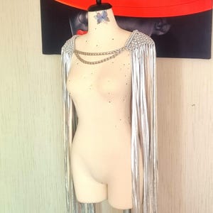 May include: A silver metallic fringe capelet featuring a textured, woven shoulder construction and two draped gold-tone metal chains connecting the front. The long, thin metallic strips hang vertically from the shoulders, creating a fluid, reflective appearance on a mannequin.