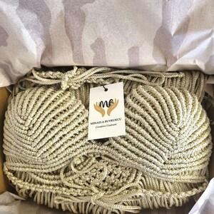May include: A gold-colored, hand-woven clutch bag with a textured, fan-like design. The bag is inside a cardboard box, lined with tissue paper. A tag with the text "MIHAELA BUYRUKCU Creative Couture" is attached to the bag.