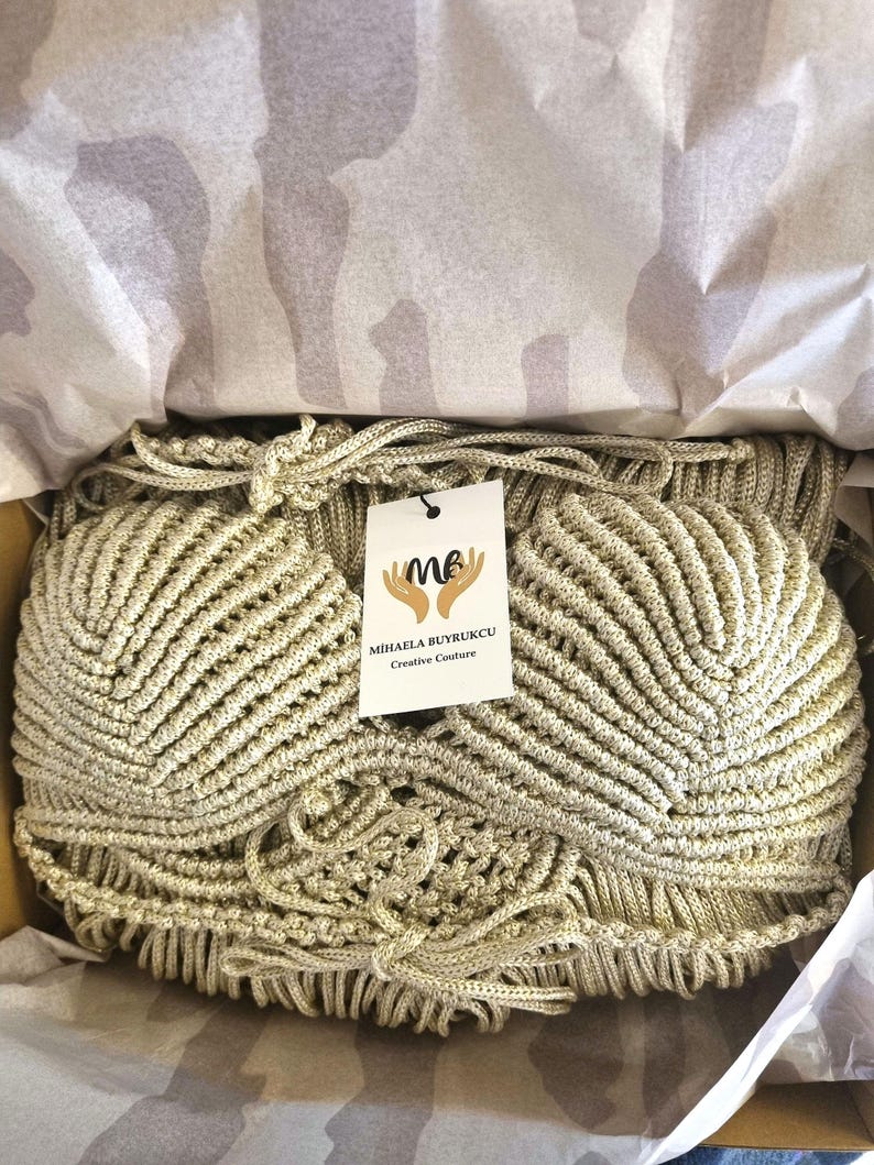 May include: A gold-colored, hand-woven clutch bag with a textured, fan-like design. The bag is inside a cardboard box, lined with tissue paper. A tag with the text "MIHAELA BUYRUKCU Creative Couture" is attached to the bag.