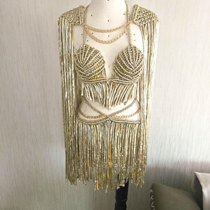 May include: A metallic gold two-piece outfit featuring a macrame-style bralette and a matching skirt with long fringe. The set includes integrated gold chain accents at the neckline and waist, along with long, flowing fringe sleeves attached to the top.