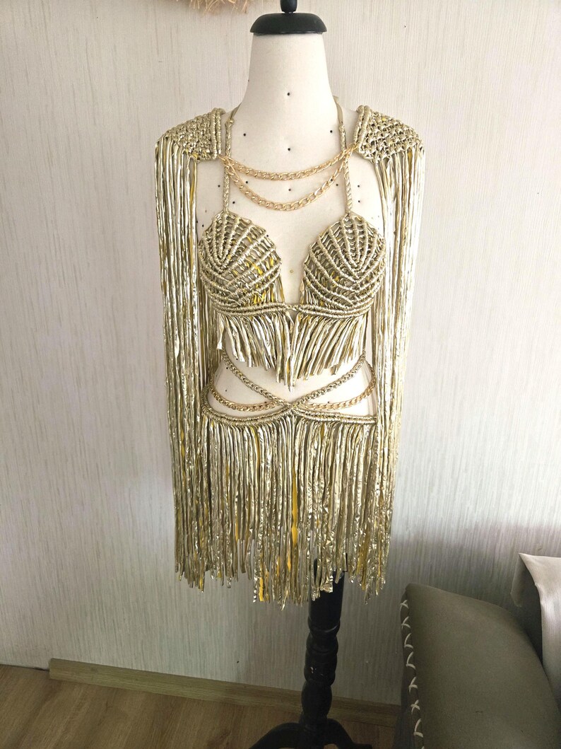 May include: A metallic gold two-piece outfit featuring a macrame-style bralette and a matching skirt with long fringe. The set includes integrated gold chain accents at the neckline and waist, along with long, flowing fringe sleeves attached to the top.