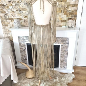 Puede incluir: Un vestido largo de macram&eacute; beige con escote halter y detalles de flecos. El vestido se muestra en un maniqu&iacute; contra una pared de piedra.
