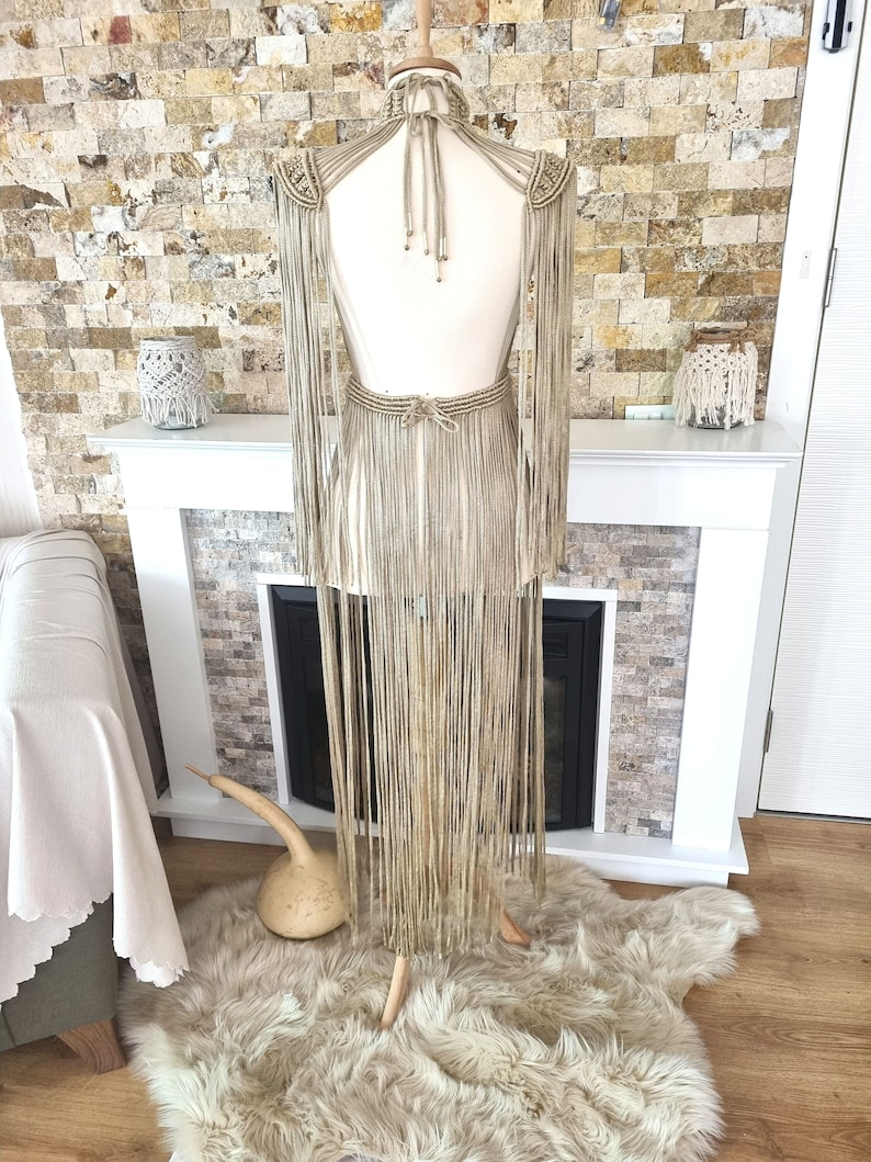 Puede incluir: Un vestido largo de macram&eacute; beige con escote halter y detalles de flecos. El vestido se muestra en un maniqu&iacute; contra una pared de piedra.