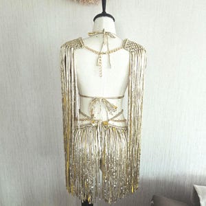 May include: A metallic gold fringe garment displayed on a mannequin, featuring long shoulder fringe panels, a back tie closure, and a waist section with multiple thin straps and dangling fringe elements that extend down to the lower back area.